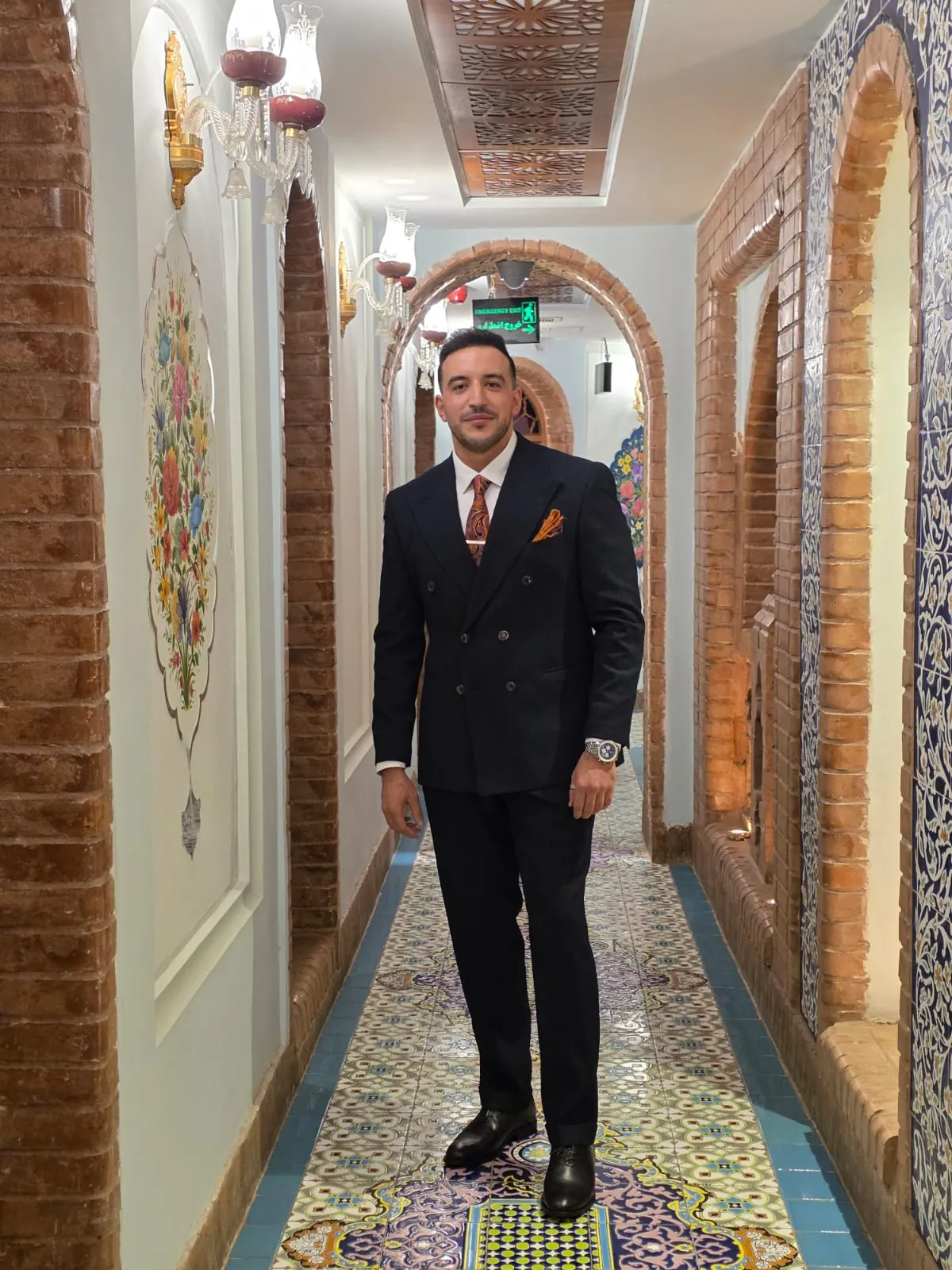Abdollah Mohammadpour in a navy double-breasted suit with orange tie, standing in an ornate hallway